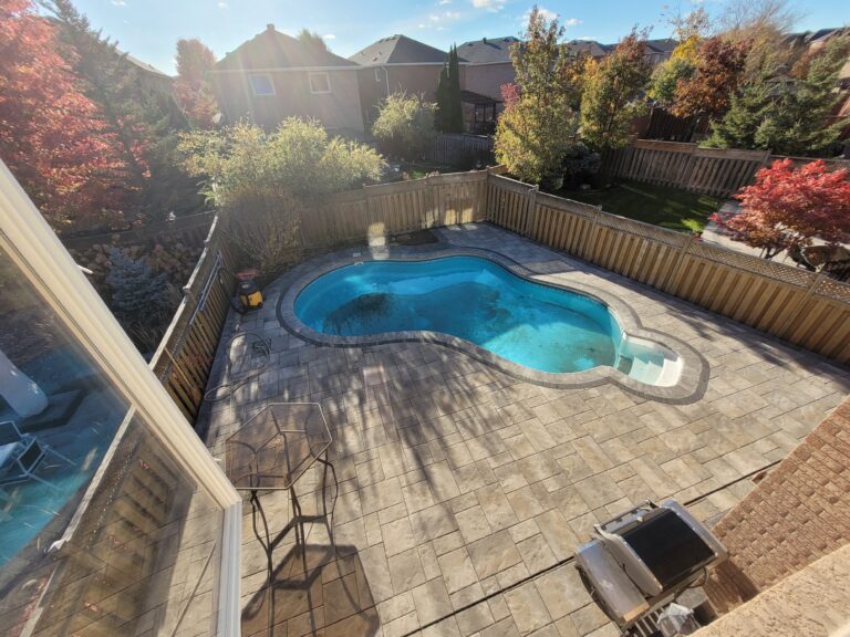 A backyard view featuring a swimming pool surrounded by a stone patio and colorful autumn foliage.